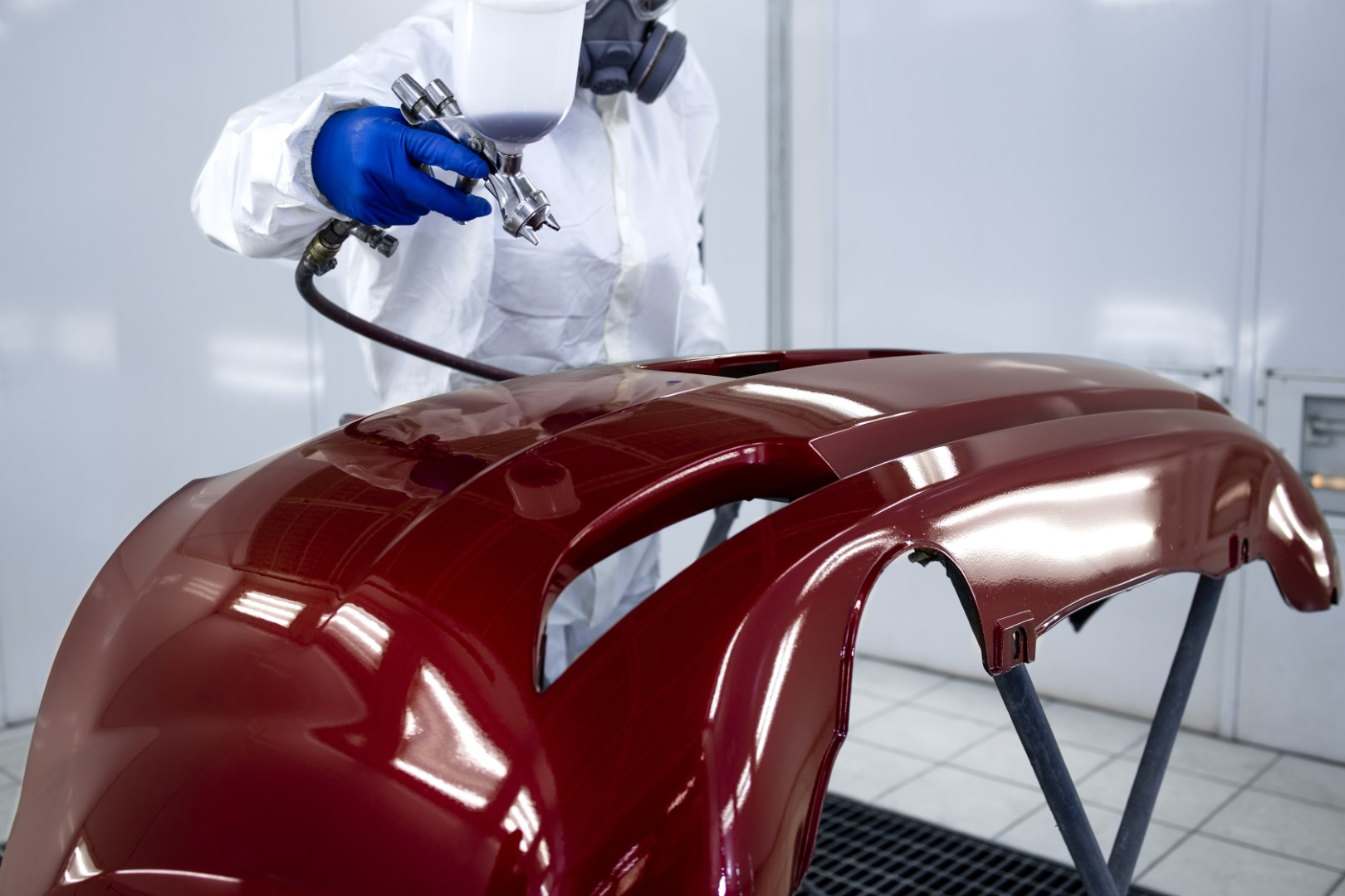 Car painter in protective clothes and mask painting automobile bumper with metallic paint and varnish in chamber workshop.